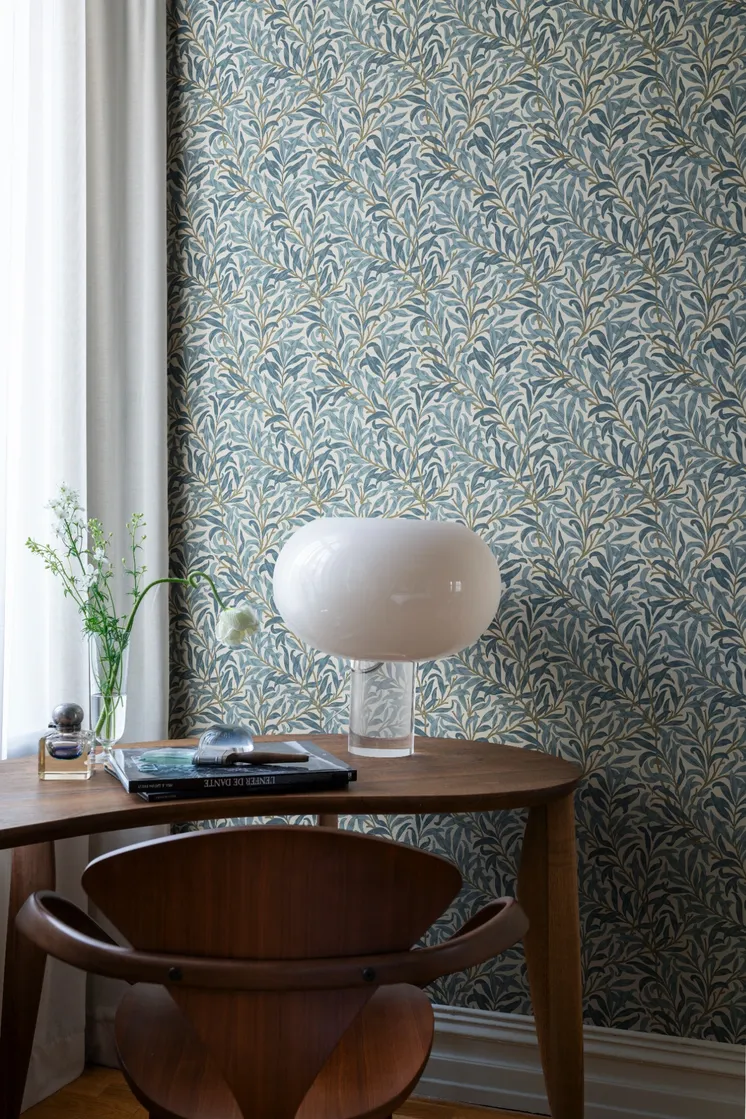 Blå bladmönstrad tapet med slingrande grenar, träbord, designlampa och dekorativa detaljer.
