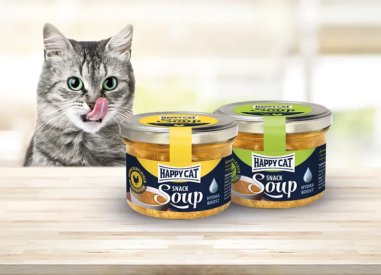 Katze sitzt vor Happy Cat Snack Soup, schleckt sich das Maul