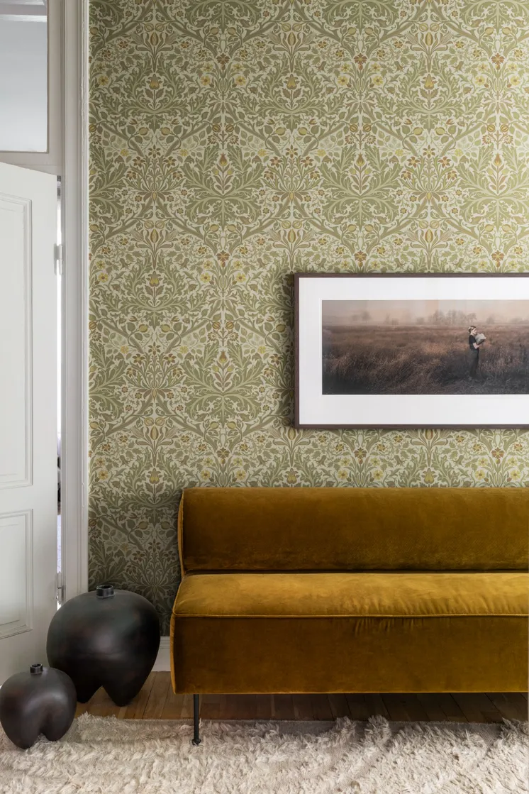 Interiör med ljus botanisk tapet i nougat, khaki och beige, senapsgul sammetssoffa och inramad tavla.