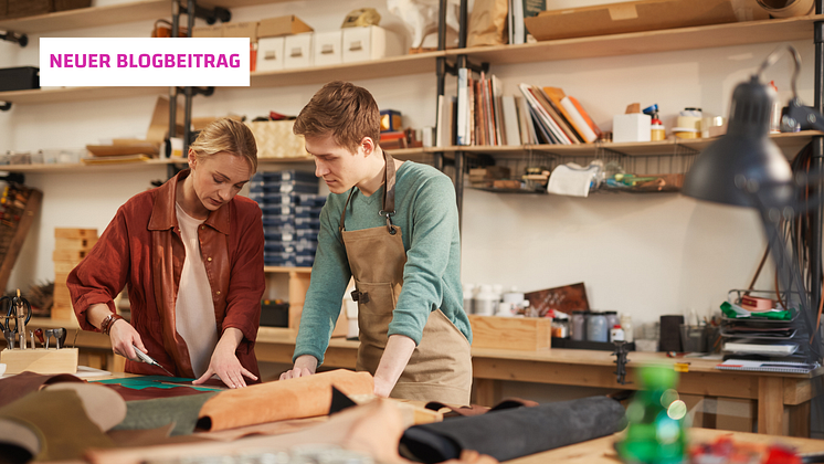 Zwei Personen in einer Werkstatt, die gemeinsam an einem Tisch mit Lederstücken arbeiten.  