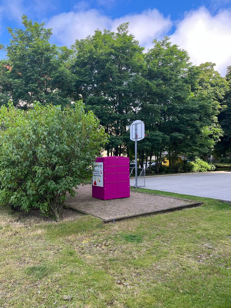 En rosa Pifflbox placerad på en grusad yta omgiven av grönska i Sorgenfri, Malmö. I bakgrunden syns träd och en mindre asfalt