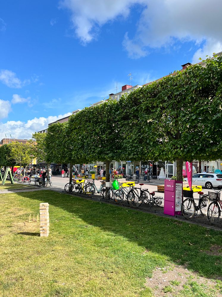 Pifflbox bredvid cyklar och spel i parkmiljö, med människor och butiker i bakgrunden i Linköpings kommun.