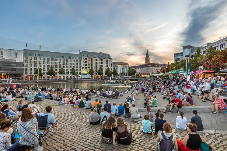 Menschen versammeln sich um den Kieler Bootshafen, um auf einer großen Leinwand die Livepremiere eines Theaterstücks zu sehen