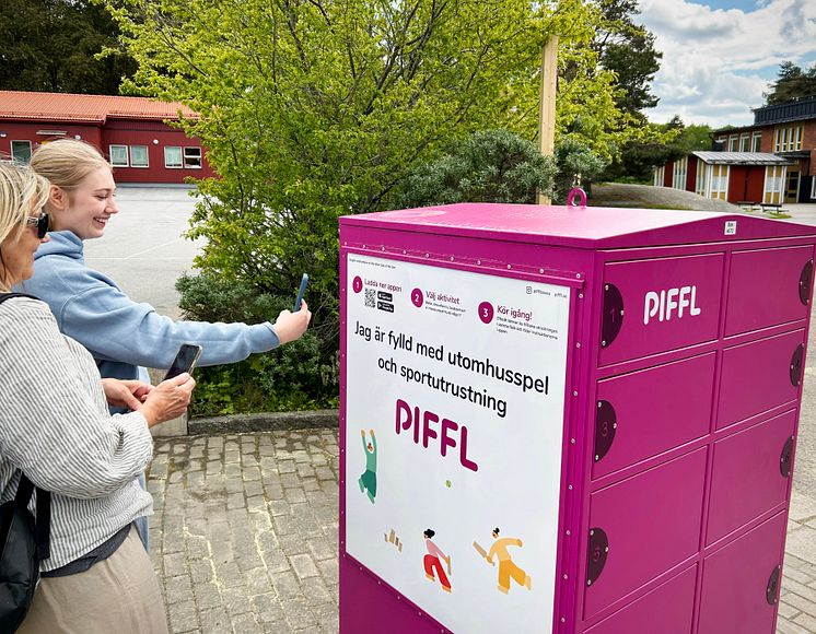 Två personer står vid en rosa Pifflbox utomhus och använder sina mobiltelefoner för att låsa upp aktiviter gratis.