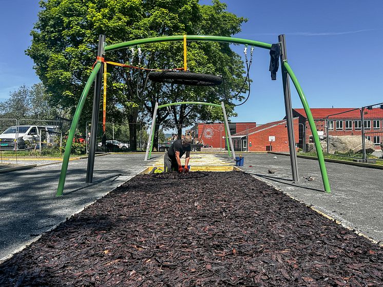 Person legger barkflis under klatrestativ med ringer på lekeplass, med blå himmel og Trasop skole i bakgrunnen.