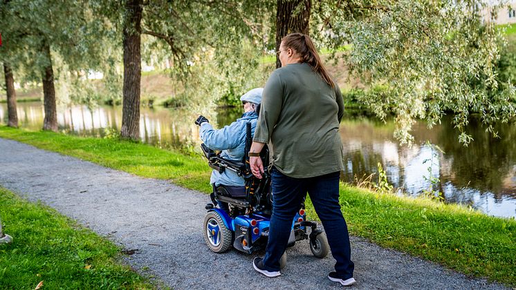 En kvinnlig assistent är ute och går med en person i rullstol i gröna omgivningar.