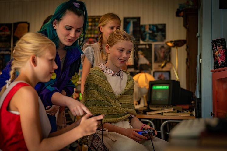 En eldre ungdom utkledd i 90-tallsklær viser tre mindre jenter hvordan de spiller på et gammelt dataspill. Foto: Anno museum