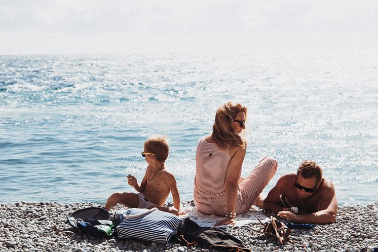 Eine Familie sitzt entspannt am Kiesstrand vor dem Meer. Eine Frau, ein Mann und ein Kind genießen die Sonne.