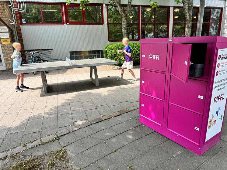 Två barn spelar pingis vid ett pingisbord intill en rosa Piffl-aktivitetsbox. Boxens skåpdörrar är öppna, redo för utlåning.