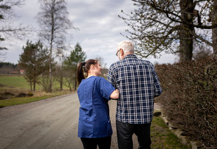 Äldre man går på skogsväg med stöd av hemtjänstpersonal