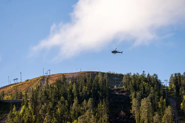 En helikopter flyger över ett grönt fjäll under blå himmel.