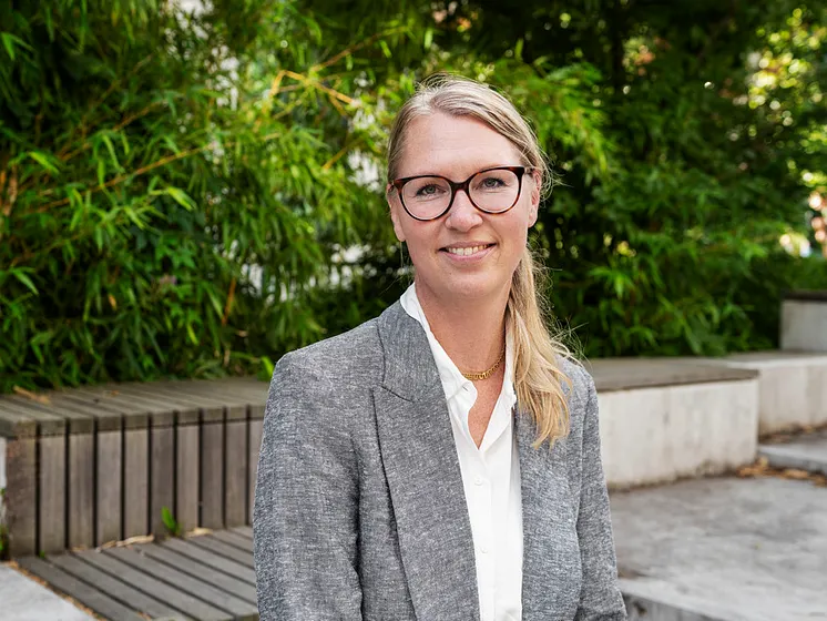 Blond kvinna med glasögon. Hon bär vit skjorta och grå kavaj. 