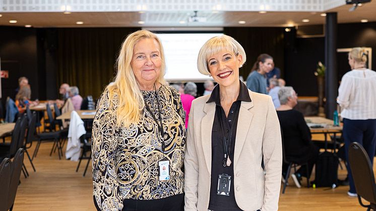 Två leende kvinnor poserar i ett stort mötesrum, i bakgrunden ses många stående och sittande deltagare på evenemnaget.