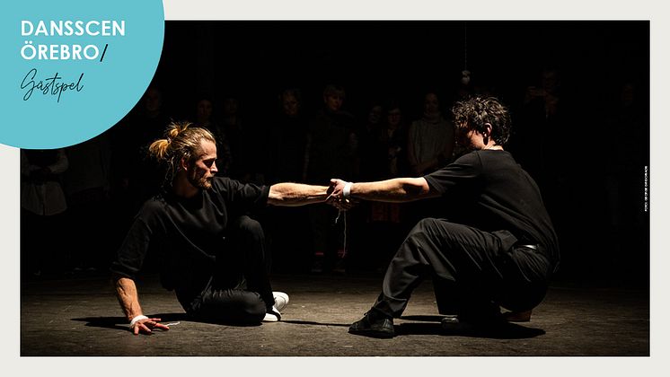 Två manliga dansare sitter på scengolv och håller i varandras utsträckta arm.