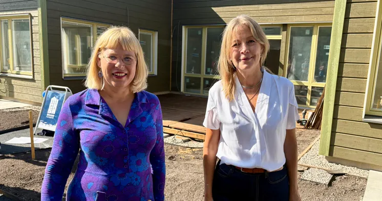 Två kvinnor står framför ett hus i grönmålat trä. I bakgrunden syns en uteplats och en skylt med Micasa Fastigheters logotyp.