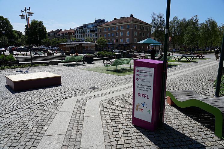 En rosa Pifflbox står i stadspark i Alingsås. Det finns picknickbord och människor som vistas i det offentliga rummet.
