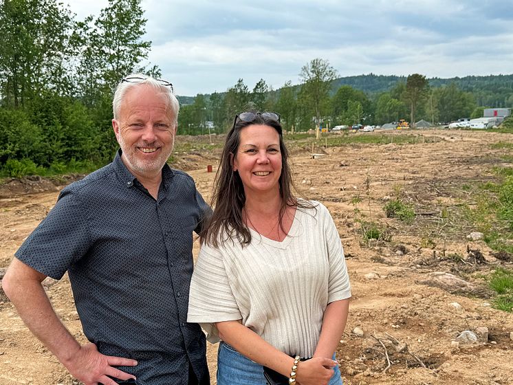 Två personer framför ett byggnadsområde där det grävs i marken. I bakgrunden syns skog och byggnader.