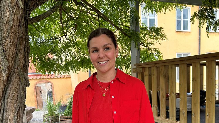 Lina Ejlertsson står under ett lövträd och ser in i kameran. Hon har på sig en röd tröja, röd skjorta och vita byxor.