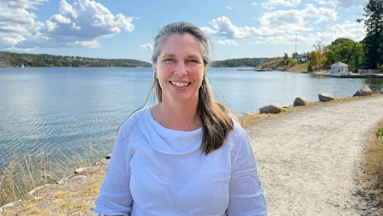 Forskare Eva Lindell har en vit blus och ler mot kameran, i bakgrunden syns utsikt över vatten och grönområden.
