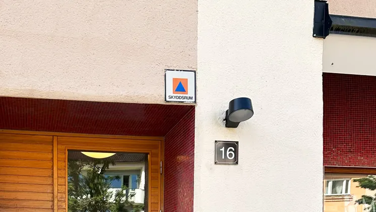 Entré till bostadshus med skyddsrumsskylt ovanför ingången och husnummer 16 på fasaden