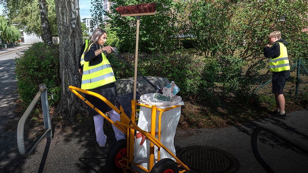 En ungdom som plockar skräp på en gata med en gul sopvagn.