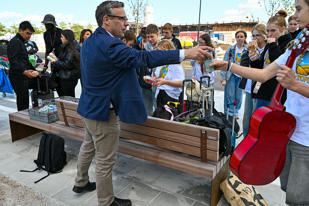 En ung kille i tonåren står bredvid en bänk och håller i en gitarr. En man räcker honom en burk kolsyrat vatten.