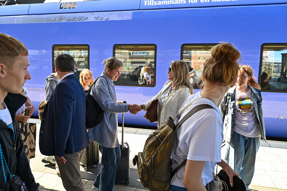 En grupp människor som står bredvid ett blått tåg. Två kvinnor tar i hand och hälsar på varandra.
